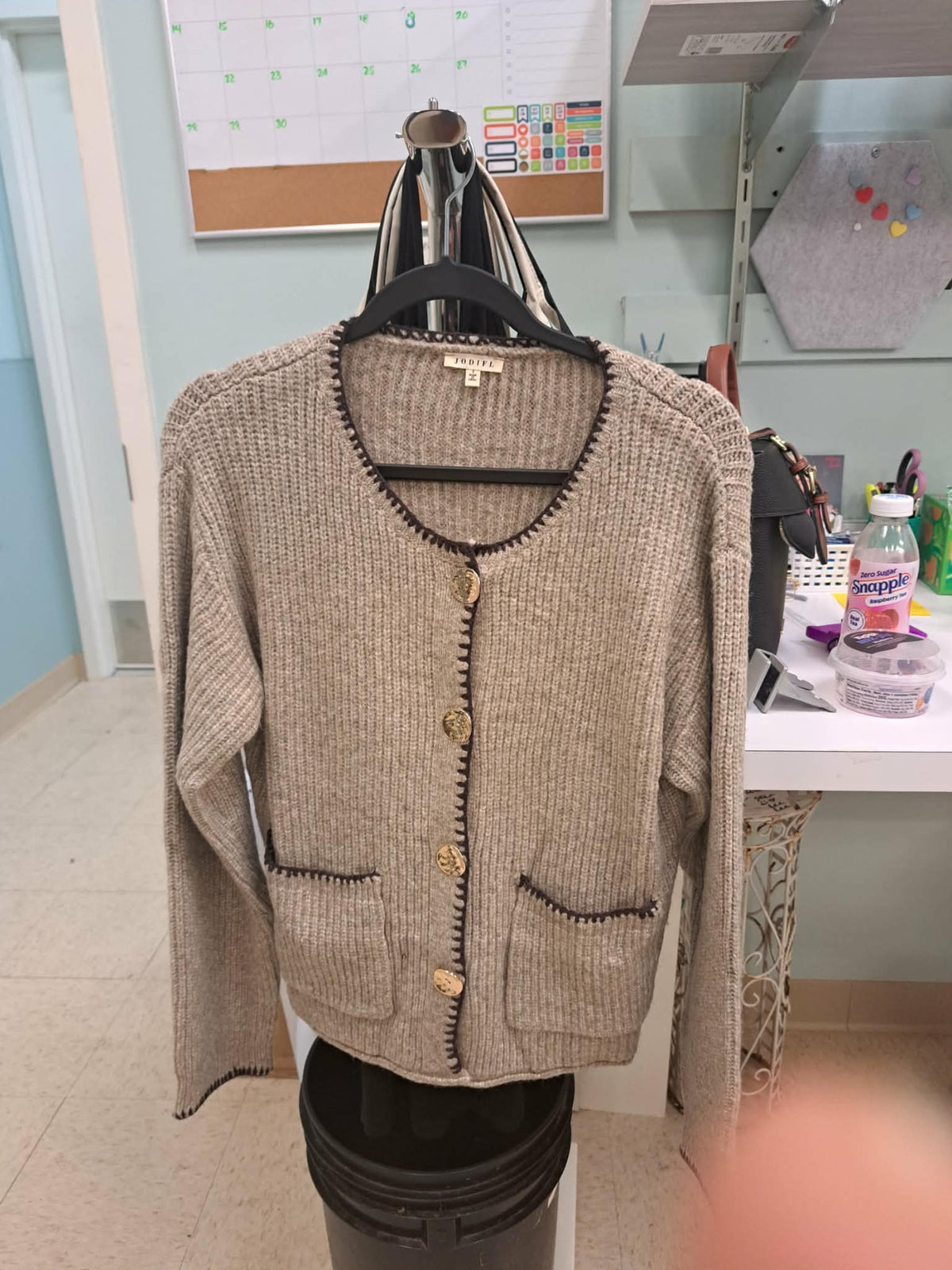 A beige ribbed knit cardigan sweater with gold buttons and black trim, displayed on a hanger on a mannequin stand in an indoor room.