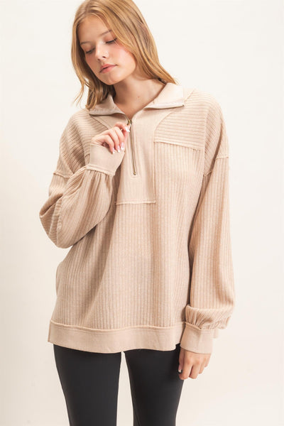 A woman wearing a beige ribbed long-sleeve quarter-zip pullover sweater with a mock neck and elastic cuffs, paired with black leggings, against a plain off-white background.