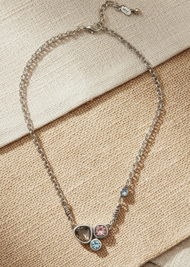A silver chain necklace featuring three small heart-shaped charms in dark gray, light gray, and rose gold, accented with tiny clear stones, shown on a light gray background.