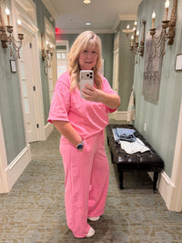 A woman takes a mirror selfie wearing a matching pink scrub top and scrub pants set in a hallway with decorative wall sconces and a bench.