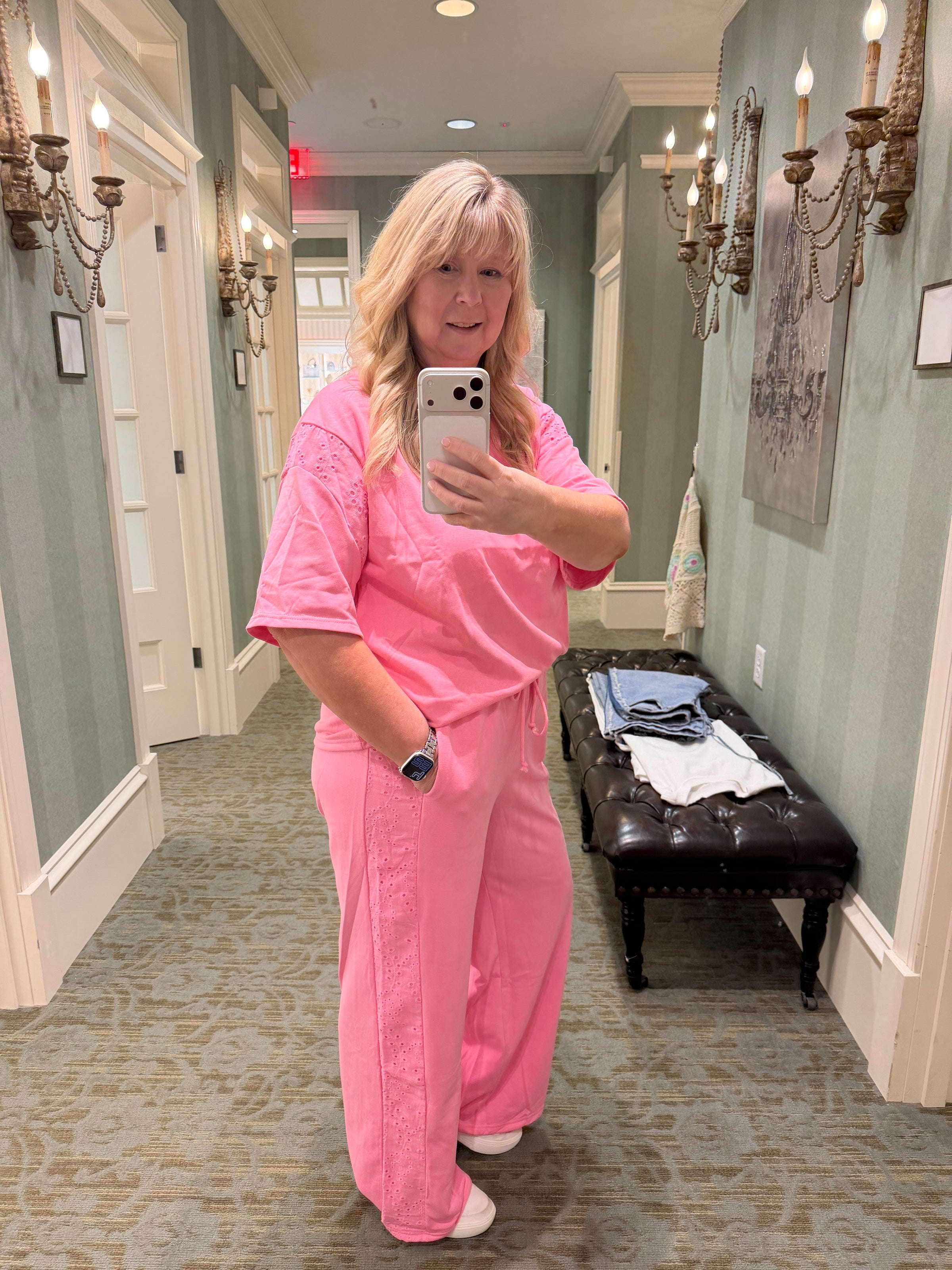 A woman takes a mirror selfie wearing a matching pink scrub top and scrub pants set in a hallway with decorative wall sconces and a bench.