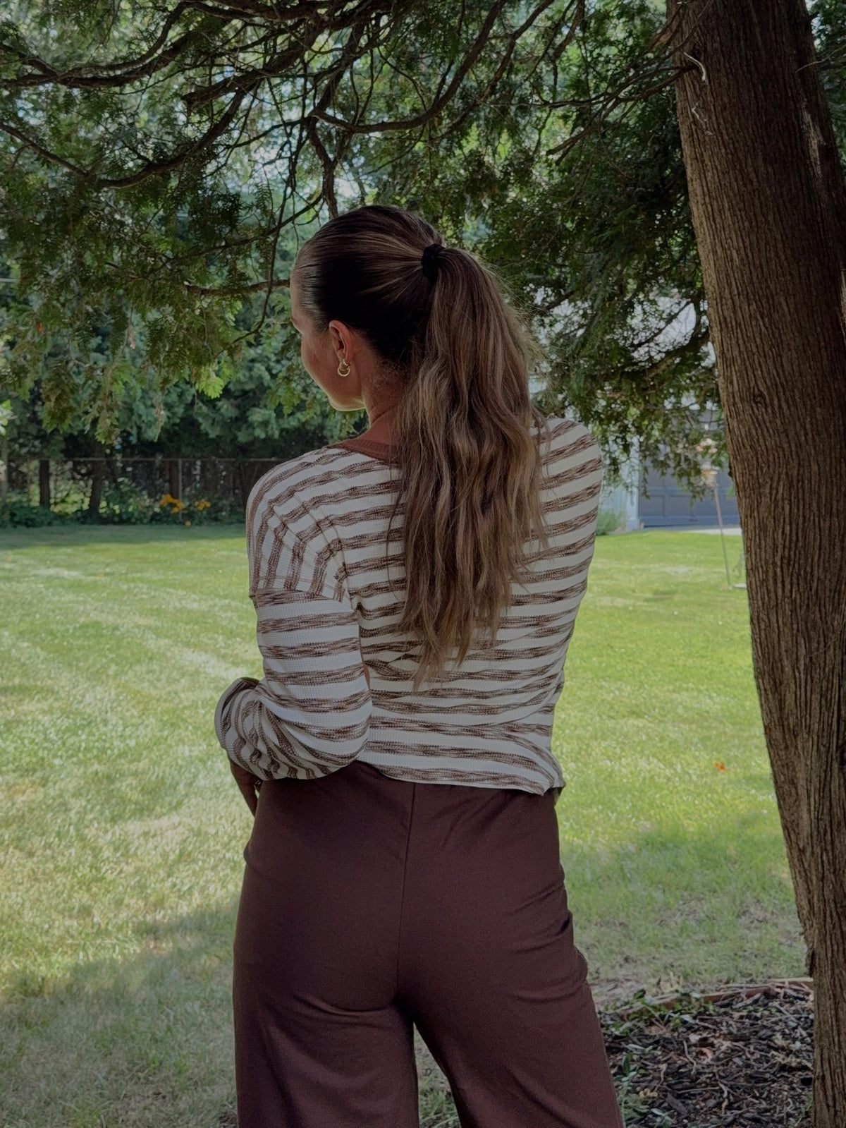 A woman viewed from behind stands in a backyard under a large tree, wearing a beige and brown horizontally striped long-sleeve top and dark brown wide-leg pants.