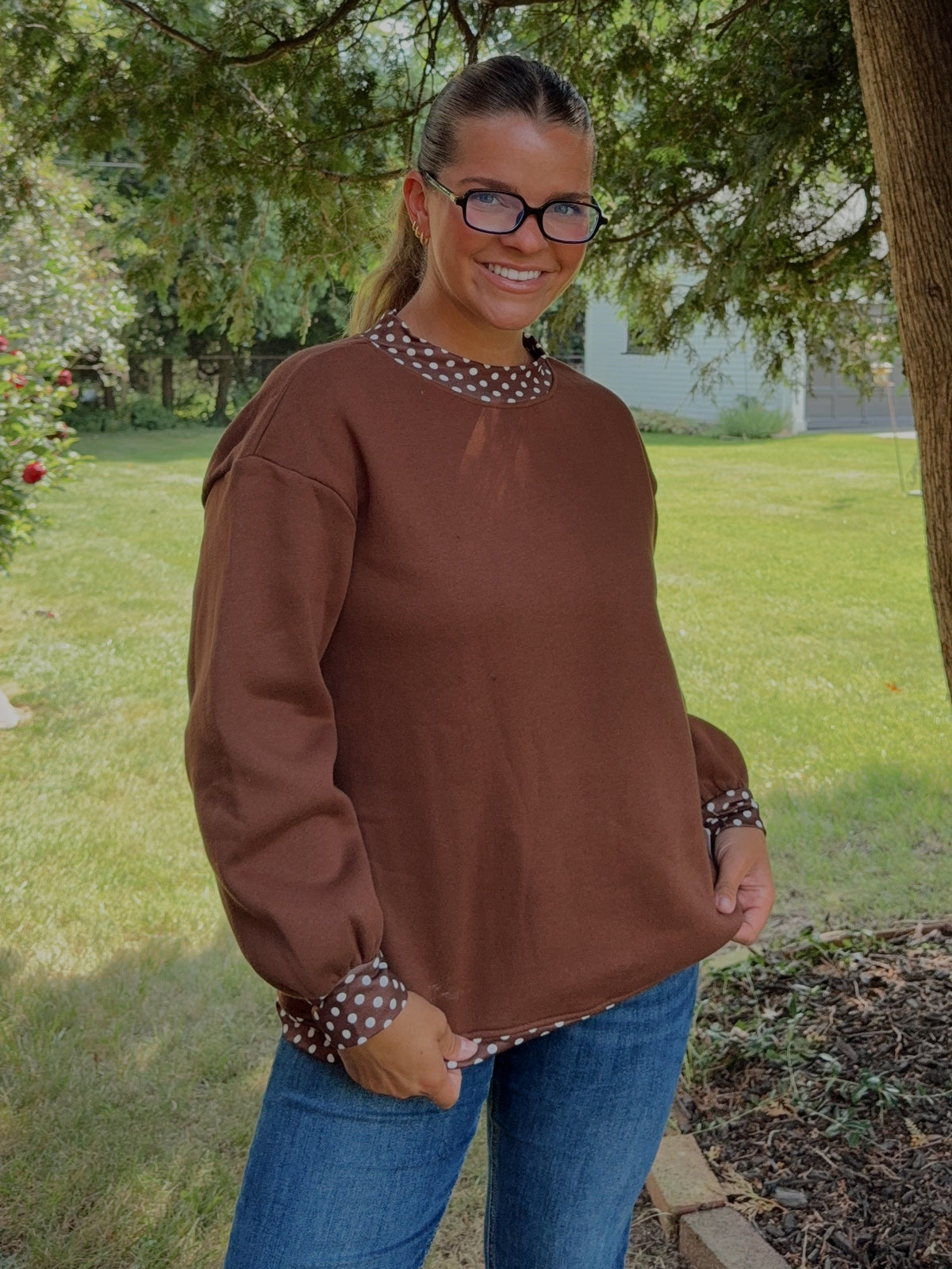 A woman wearing a brown long-sleeve top with patterned trim and blue jeans, standing outdoors under a large tree in a grassy yard.