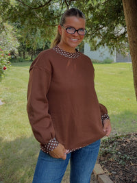 A woman wearing a brown long-sleeve top with patterned trim and blue jeans, standing outdoors under a large tree in a grassy yard.