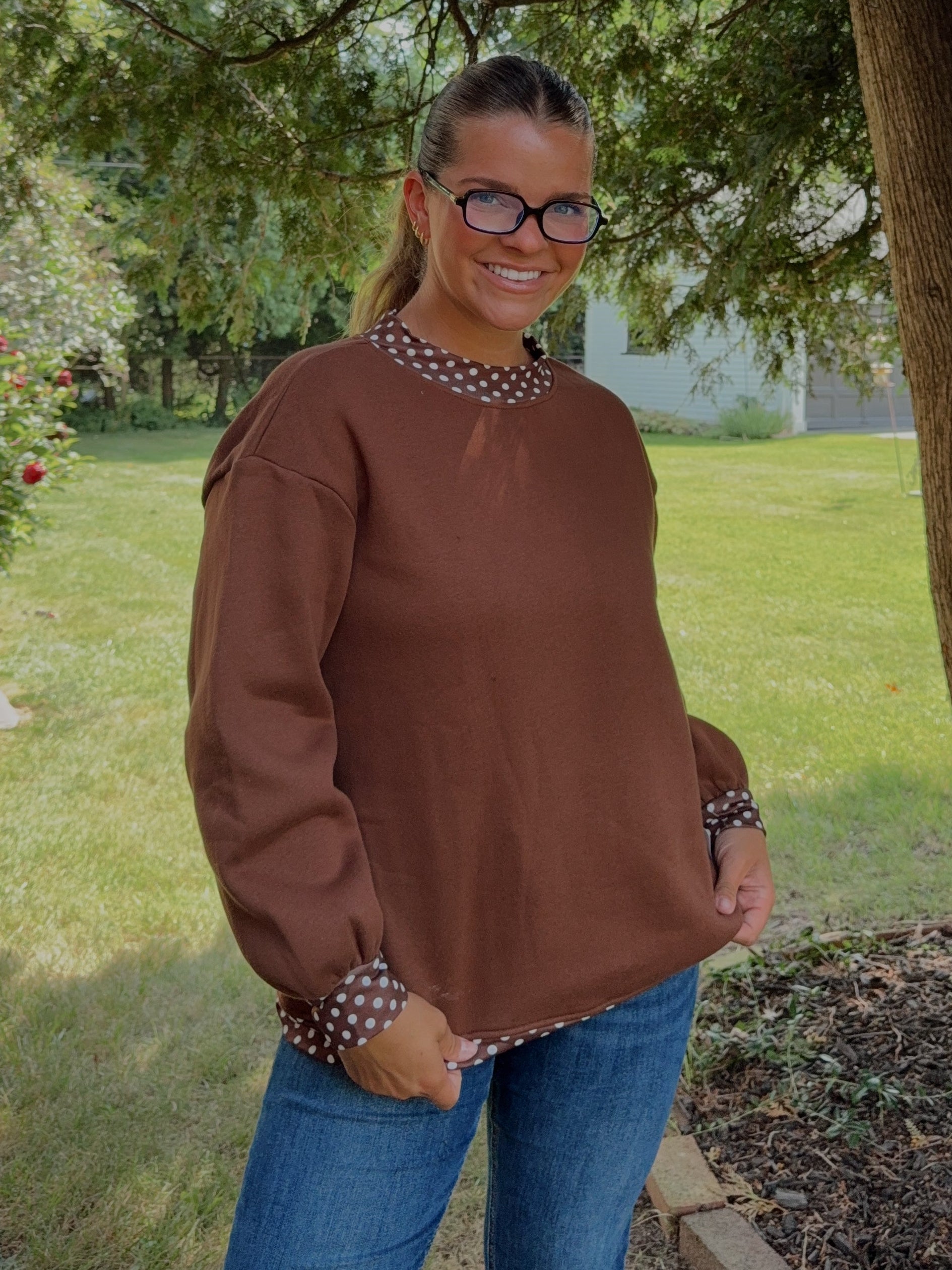 A woman wearing a brown long-sleeve top with patterned trim and blue jeans, standing outdoors under a large tree in a grassy yard.