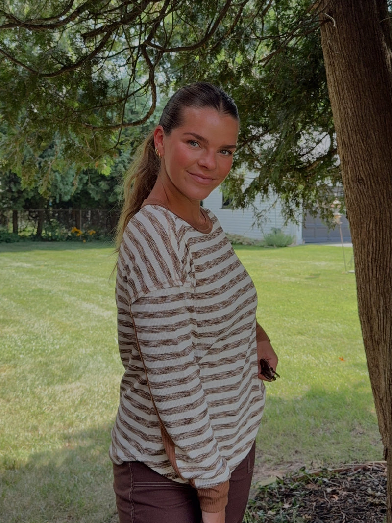A woman standing outdoors in a grassy yard next to a large tree trunk, wearing a beige and brown horizontally striped long-sleeve top and brown pants.